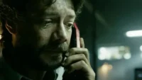 Homem barbudo de cabelo escuro, com lágrima, ouve ao telefone vermelho. Ambiente escuro e tenso. Clima dramático.
