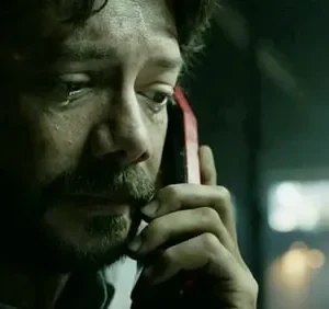Homem barbudo de cabelo escuro, com lágrima, ouve ao telefone vermelho. Ambiente escuro e tenso. Clima dramático.