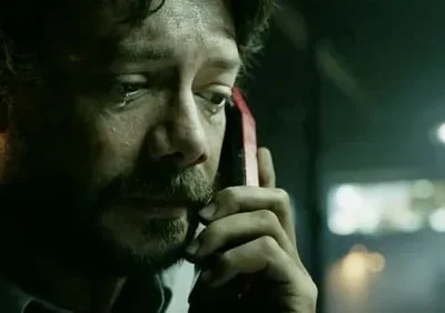 Homem barbudo de cabelo escuro, com lágrima, ouve ao telefone vermelho. Ambiente escuro e tenso. Clima dramático.