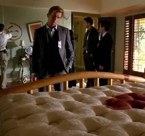 Um homem de terno observa uma cama ensanguentada num quarto. Na parede, um grande rosto sorridente vermelho. Outros investigadores ao fundo.