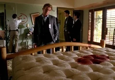 Um homem de terno observa uma cama ensanguentada num quarto. Na parede, um grande rosto sorridente vermelho. Outros investigadores ao fundo.