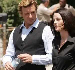 Homem loiro de colete listrado e mulher morena andam lado a lado em rua ensolarada. No fundo, pessoas desfocadas interagem.