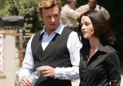 Homem loiro de colete listrado e mulher morena andam lado a lado em rua ensolarada. No fundo, pessoas desfocadas interagem.