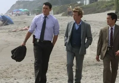 Três homens formalmente vestidos, dois com distintivos, caminham numa praia ensolarada. Pessoas e guarda-sóis ao fundo. Atmosfera de investigação.