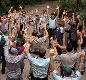 Multidão de pessoas, incluindo oficiais, com as mãos levantadas na floresta. Clima de rendição ou confronto.