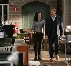 Homem de terno e mulher com distintivo caminham por um escritório movimentado. Outras pessoas trabalham. Ambiente ativo e investigativo.