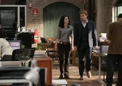 Homem de terno e mulher com distintivo caminham por um escritório movimentado. Outras pessoas trabalham. Ambiente ativo e investigativo.