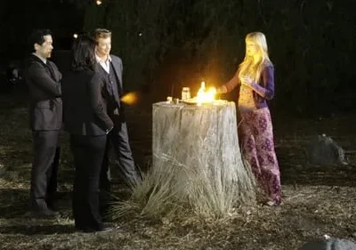Noite: mulher loira de roxo com fogo em toco. Três pessoas de terno observam em clareira. Clima misterioso.