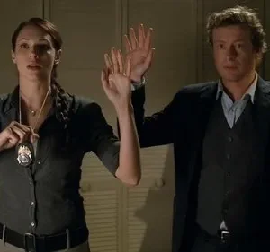 Mulher de camisa escura com distintivo e homem de terno levantam as mãos. Ambos sérios, em ambiente fechado.