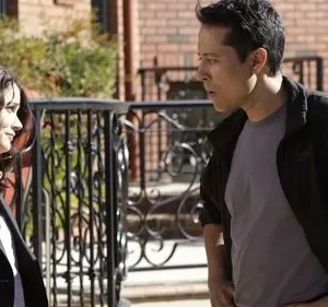 Mulher sorri para homem com expressão surpresa. Cenário urbano com tijolos e grade preta. Dia ensolarado.