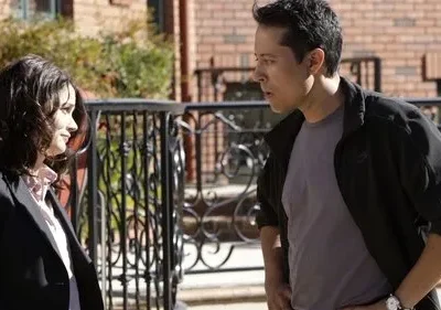 Mulher sorri para homem com expressão surpresa. Cenário urbano com tijolos e grade preta. Dia ensolarado.