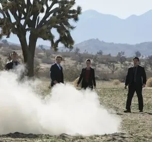 Quatro pessoas, três de terno, observam fumaça branca saindo do chão em deserto com montanhas e uma árvore de Josué.