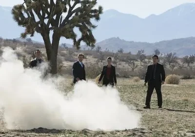 Quatro pessoas, três de terno, observam fumaça branca saindo do chão em deserto com montanhas e uma árvore de Josué.