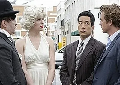 Homem como Chaplin e mulher como Marilyn Monroe parados em rua. Dois homens de terno os observam. Clima intrigante.