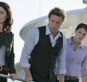 Três personagens sérios (duas mulheres, um homem) num iate. Todos olham para baixo. Uma mulher usa distintivo na cintura.