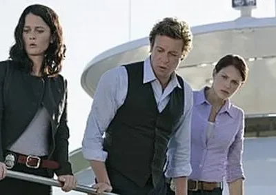 Três personagens sérios (duas mulheres, um homem) num iate. Todos olham para baixo. Uma mulher usa distintivo na cintura.