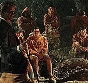 Homem de terno guia jovens de hoodies laranja em reunião noturna na floresta. Clima sério, quase ritualístico, sob luz fraca.