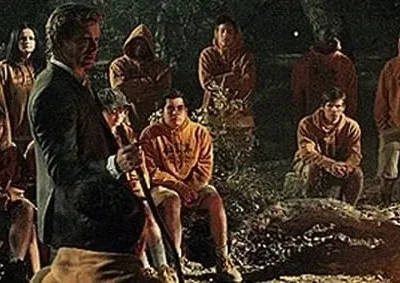 Homem de terno guia jovens de hoodies laranja em reunião noturna na floresta. Clima sério, quase ritualístico, sob luz fraca.