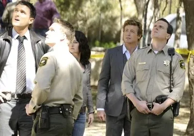 Um grupo de homens, incluindo um xerife e detetives, olham para cima em uma área externa isolada com fita amarela. Ambiente de mistério.