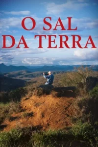 Homem de chapéu ajoelhado em morro de terra, fotografando vastas montanhas sob céu azul e nuvens brancas.