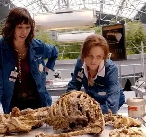 Duas cientistas em jalecos azuis examinam um esqueleto em um laboratório moderno com teto de vidro. Clima investigativo.