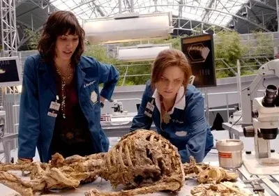 Duas cientistas em jalecos azuis examinam um esqueleto em um laboratório moderno com teto de vidro. Clima investigativo.