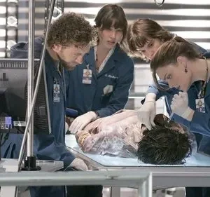 Cinco investigadores em jalecos azuis examinam um corpo em uma mesa de laboratório forense. Clima sério de análise científica.