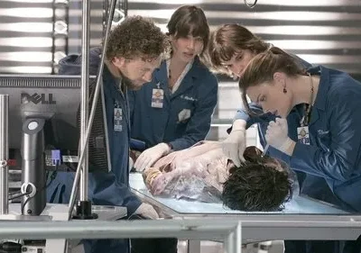 Cinco investigadores em jalecos azuis examinam um corpo em uma mesa de laboratório forense. Clima sério de análise científica.