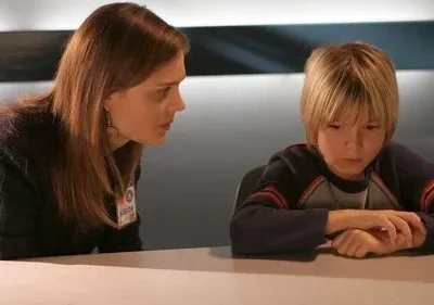 Mulher observa menino loiro sentado à mesa, que olha para baixo. Ambiente sério e introspectivo.