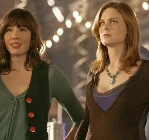 Duas mulheres em destaque. Uma sorri, com franja e mão levantada. A outra, séria, de colar. Fundo com luzes desfocadas.