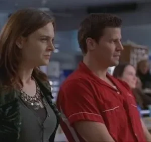 Mulher de cabelo castanho e homem de camisa de boliche vermelha. Ambos olham atentamente para a direita em uma pista de boliche.