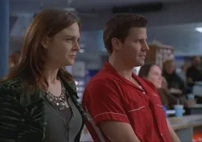 Mulher de cabelo castanho e homem de camisa de boliche vermelha. Ambos olham atentamente para a direita em uma pista de boliche.