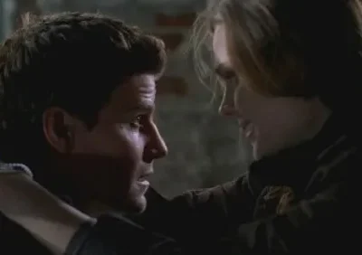 Homem com braço amarrado, com olhar apreensivo, é encarado por mulher com sorriso irônico e dominador. Cenário escuro e tenso.