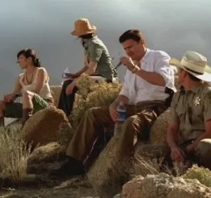 Quatro pessoas em roupas de aventura descansam em rochas e arbustos, olhando para longe em paisagem árida sob céu cinzento.