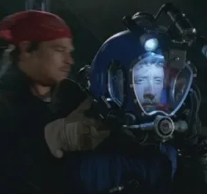 Homem de bandana assiste a outro com capacete de mergulho azul e luzes, revelando um rosto tenso na noite escura.