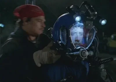 Homem de bandana assiste a outro com capacete de mergulho azul e luzes, revelando um rosto tenso na noite escura.