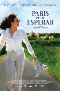Mulher sorridente com óculos de sol,cabelos ao vento,andando descalça numa estrada rural,segurando sapatos,com uma vila europeia e campos verdes ao fundo.