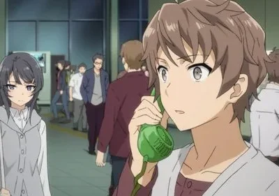 Um rapaz confuso fala ao telefone verde. Uma jovem séria com cabelos longos está por perto, no meio de uma multidão em ambiente público.