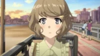 Jovem anime com cabelo castanho e olhos grandes olhando para frente, bochechas coradas, em um calçadão à beira-mar ao pôr do sol.