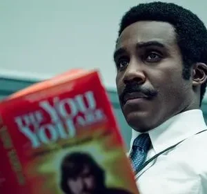Homem negro de bigode, em camisa e gravata, olha pensativo enquanto segura um livro vermelho escrito THE YOU ARE.