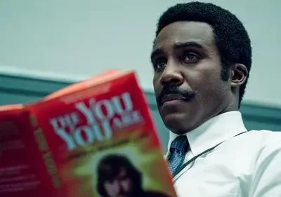 Homem negro de bigode, em camisa e gravata, olha pensativo enquanto segura um livro vermelho escrito THE YOU ARE.