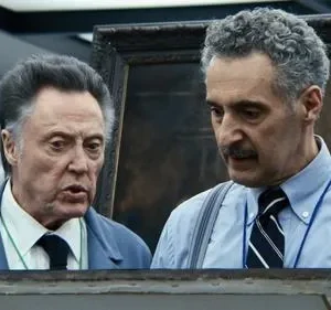 Dois homens idosos com expressões sérias examinam algo fora do quadro. Um de paletó azul, outro de camisa e suspensórios. Fundo com pintura escura.