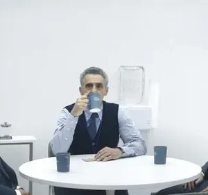 Três executivos em ternos escuros numa sala branca minimalista. Um bebe, outro observa, e o terceiro encara. Ambiente tenso.