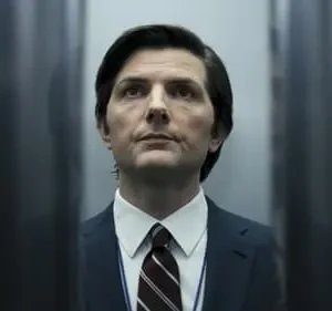 Um homem de terno sério, com cabelo escuro, enquadrado em um corredor estreito e escuro. Seu olhar é fixo, criando um clima misterioso.