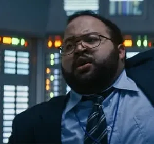 Homem barbudo de óculos e terno azul, com expressão séria, em sala de controle com muitos painéis e luzes coloridas.