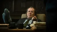 Homem de óculos, cabelo comprido, pés em meias furadas sobre mesa de escritório escuro. Gesticula, com ar cansado e descontraído.