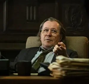 Homem de óculos, cabelo comprido, pés em meias furadas sobre mesa de escritório escuro. Gesticula, com ar cansado e descontraído.