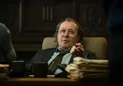 Homem de óculos, cabelo comprido, pés em meias furadas sobre mesa de escritório escuro. Gesticula, com ar cansado e descontraído.