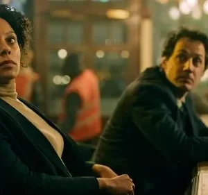 Mulher de cabelo cacheado e expressão pensativa em bar escuro. Homem ao fundo, sério, sentado ao balcão. Clima misterioso.