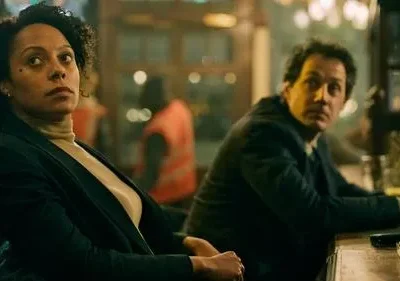 Mulher de cabelo cacheado e expressão pensativa em bar escuro. Homem ao fundo, sério, sentado ao balcão. Clima misterioso.